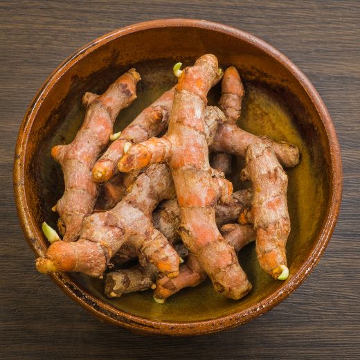 Top-down view of fresh, raw turmeric roots with small green sprouts inside a rustic clay bowl on a dark wooden table.