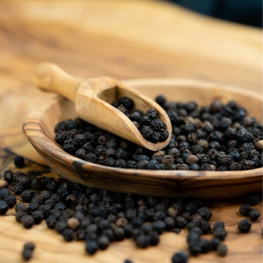 Close-up of whole black peppercorns filled inside a small wooden scoop and a rustic wooden bowl on a cutting board.