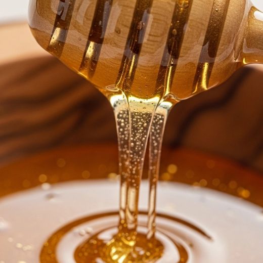 Close-up of thick, golden honey dripping from a wooden honey dipper into a bowl, showing air bubbles and a smooth, reflective texture.